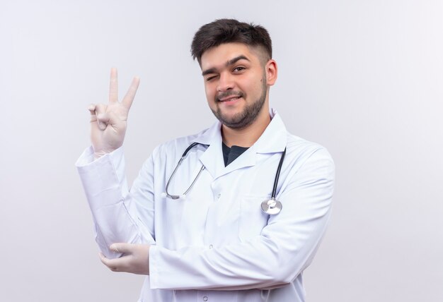 Młody przystojny lekarz w białej sukni medycznej białe rękawiczki medyczne i stetoskop mrugający jednym okiem pokazujący znak pokoju z palcami stojącymi nad białą ścianą