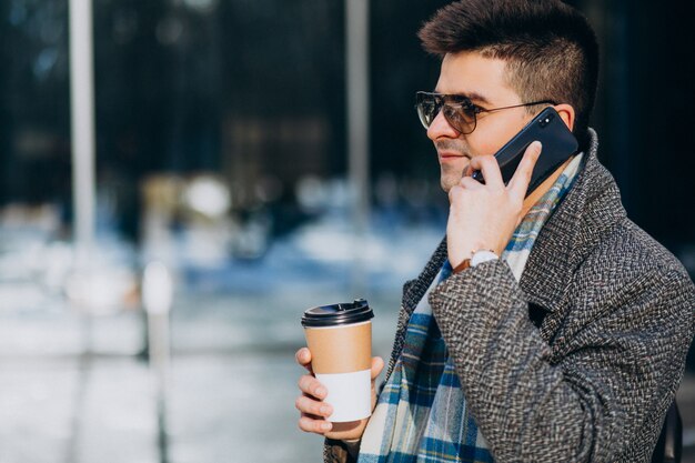 Młody przystojny mężczyzna pije kawę na zewnątrz i używa telefon