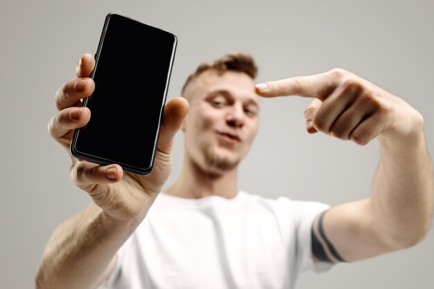 Młody przystojny mężczyzna pokazuje ekran smartfona na szarej przestrzeni z twarzą niespodzianki