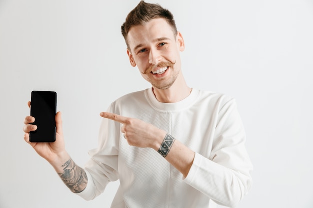 Młody przystojny mężczyzna pokazuje ekran smartfona na szarej przestrzeni z twarzą niespodzianki