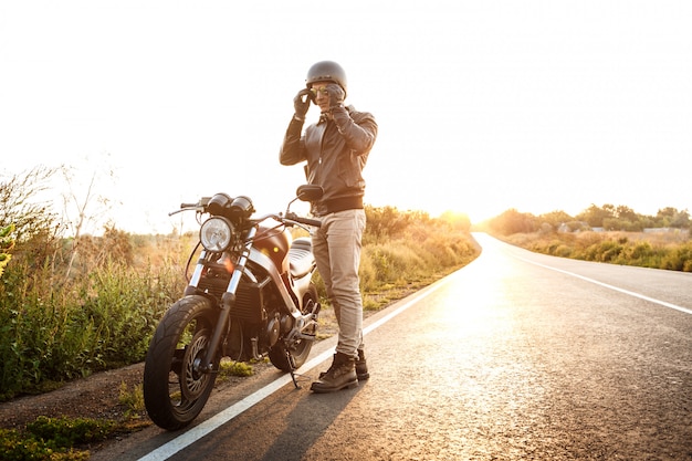 Młody przystojny mężczyzna pozuje blisko jego motocyklu przy wsi drogą.