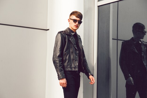 Młody przystojny mężczyzna pozuje w pobliżu nowoczesnego centrum biznesowego, ubrany w stylową skórzaną kurtkę z kolcami, czarne dżinsy i okulary przeciwsłoneczne, brutalny wygląd.