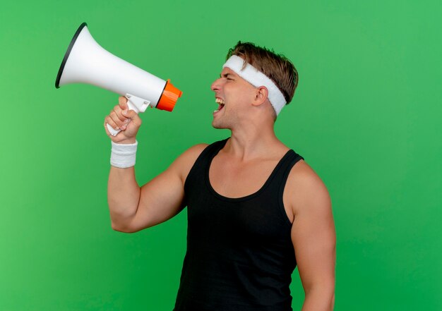 Młody przystojny mężczyzna sportowy noszenie opaski i opaski na rękę krzycząc w głośniku na białym tle na zielonym tle z miejsca na kopię