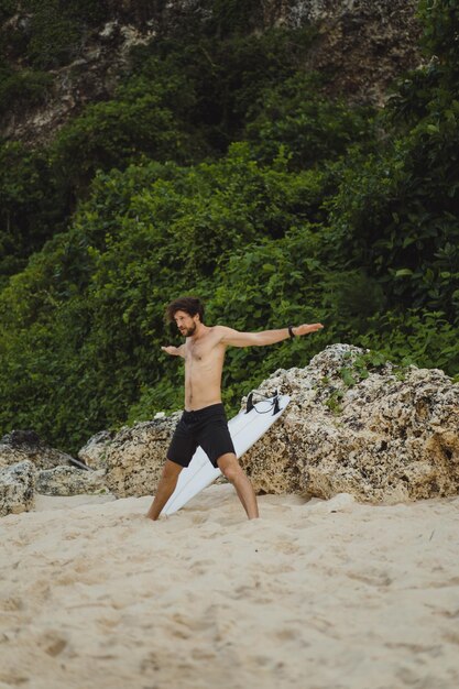 Młody przystojny mężczyzna surfer na brzegu oceanu robi rozgrzewkę przed surfowaniem. ćwiczenia przed sportem, stretching przed surfingiem.