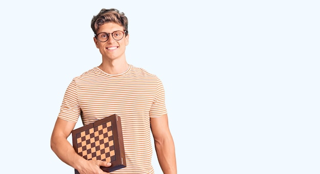 Bezpłatne zdjęcie młody przystojny mężczyzna trzymający szachy, wyglądający pozytywnie i szczęśliwy, stojąc i uśmiechając się z pewnym uśmiechem pokazującym zęby