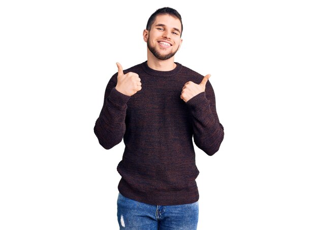 Młody przystojny mężczyzna ubrany w swobodny sweter ze znakiem sukcesu, wykonujący pozytywny gest z kciukami w górę, uśmiechnięty i szczęśliwy, wesoły wyraz twarzy i gest zwycięzcy