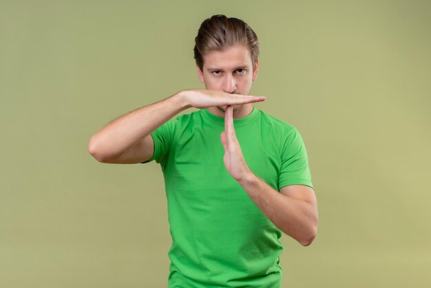 Młody przystojny mężczyzna ubrany w zieloną koszulkę z marszczoną twarzą, robiąc gest z rękami stojącymi na zielonej ścianie