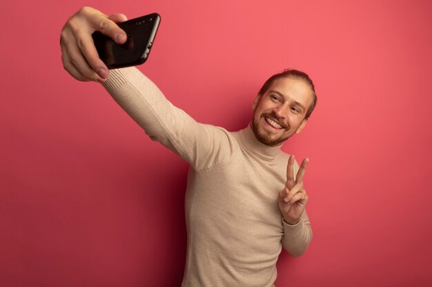 Młody przystojny mężczyzna w beżowym golfie za pomocą smartfona robi selfie, uśmiechając się, pokazując znak v