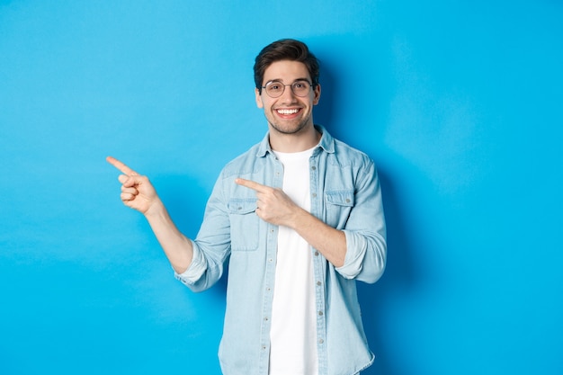 Młody przystojny mężczyzna w okularach pokazujący reklamę, uśmiechający się i wskazujący palcami w lewo, ogłaszający ogłoszenie, stojący na niebieskim tle