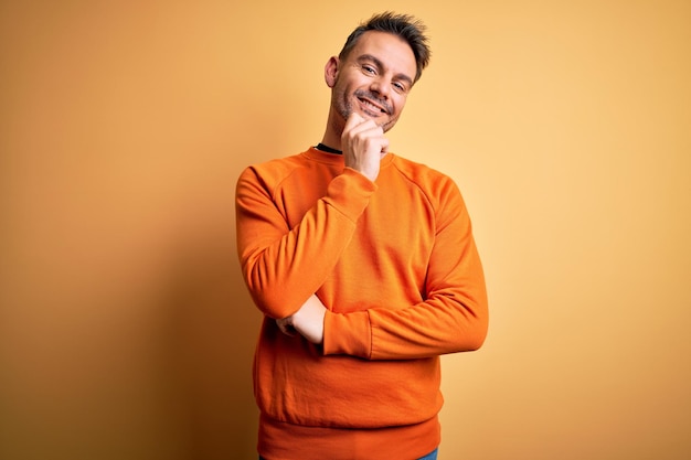 Bezpłatne zdjęcie młody przystojny mężczyzna w pomarańczowym swobodnym swetrze, stojący nad odizolowanym żółtym tłem, patrzący pewnie w kamerę z uśmiechem ze skrzyżowanymi ramionami i ręką uniesioną na brodzie pozytywne myślenie