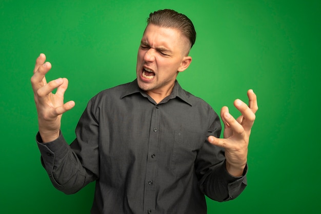 Bezpłatne zdjęcie młody przystojny mężczyzna w szarej koszuli krzyczy i krzyczy z wyciągniętymi ramionami szalony i zły, stojąc nad zieloną ścianą