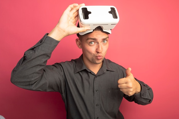 Bezpłatne zdjęcie młody przystojny mężczyzna w szarej koszuli w okularach wirtualnej rzeczywistości, pewny siebie, pokazując kciuki do góry stojąc nad różową ścianą