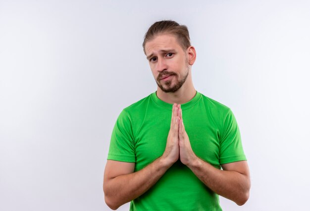 Młody przystojny mężczyzna w zielonej koszulce błagając i modląc się z rękami razem emocjonalny i zmartwiony stojąc na białym tle