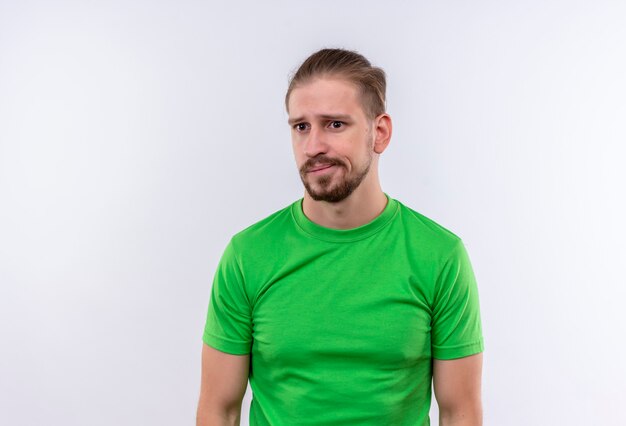 Młody przystojny mężczyzna w zielonej koszulce wyglądający na zdezorientowanego i bardzo niespokojnego stojącego na białym tle