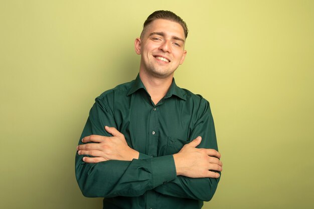 Młody przystojny mężczyzna w zielonej koszuli z szczęśliwą twarzą ze skrzyżowanymi rękami na piersi, uśmiechając się radośnie