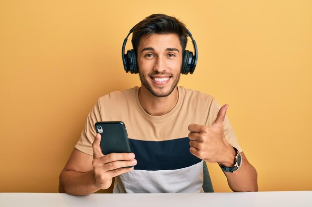 Młody przystojny mężczyzna za pomocą smartfona w słuchawkach uśmiechnięty szczęśliwy i pozytywny, kciuk w górę robi doskonały i znak aprobaty