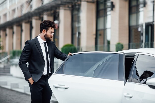 Młody przystojny odnoszący sukcesy menedżer w czarnym garniturze wsiadający na tylne siedzenie swojego samochodu w pobliżu nowoczesnego centrum biznesowego, na ulicy wielkiego miasta