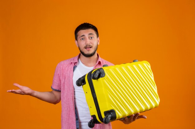 Młody przystojny podróżnik facet trzyma walizkę, nieświadomy i zdezorientowany, patrząc na aparat stojący z podniesionym ramieniem, mający wątpliwości na pomarańczowym tle