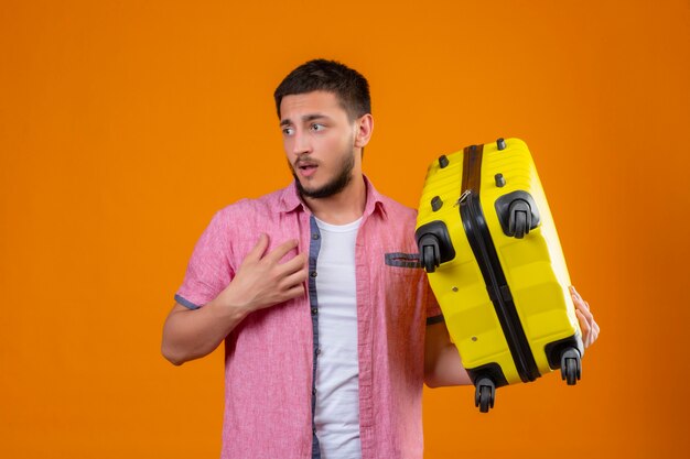 Młody przystojny podróżnik facet trzyma walizkę patrząc na bok ze smutnym wyrazem twarzy niezadowolony stojąc na pomarańczowym tle
