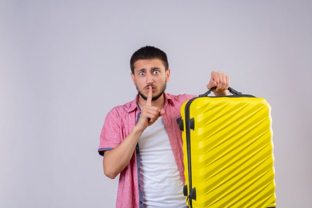 Młody przystojny podróżnik facet trzyma walizkę robi ciszy gest z palcem na ustach patrząc na aparat zaskoczony stojąc na białym tle