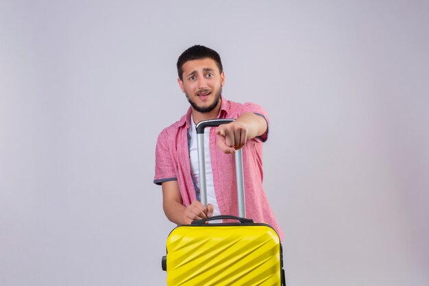 Młody przystojny podróżnik facet trzyma walizkę wskazując palcem na aparat ze smutnym wyrazem twarzy nieszczęśliwej i niezadowolonej stojącej na białym tle