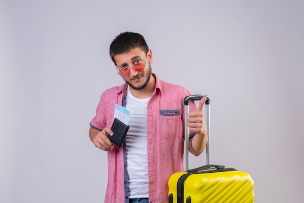 Młody przystojny podróżnik facet w okularach przeciwsłonecznych, trzymając walizkę i bilety lotnicze, patrząc na kamery z pewnym uśmiechem pokazując kciuki do góry stojąc na białym tle