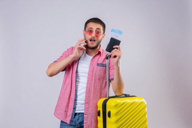 Młody przystojny podróżnik facet w okularach przeciwsłonecznych, trzymając walizkę i bilety lotnicze, patrząc zdezorientowany, mając wątpliwości stojąc na białym tle