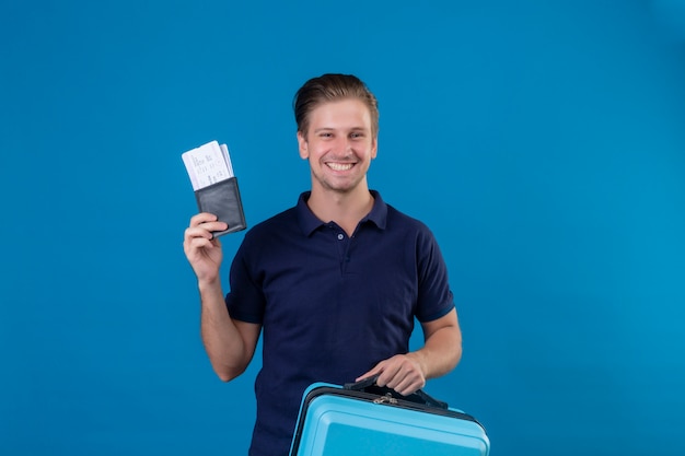 Młody Przystojny Podróżnik Mężczyzna Posiadający Bilety Lotnicze Szczęśliwy I Pozytywny Patrząc Na Kamery Z Uśmiechem Na Twarzy Stojącej Na Niebieskim Tle
