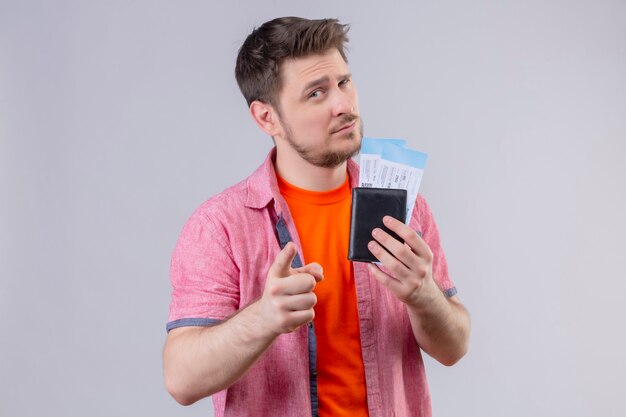 Młody przystojny podróżnik mężczyzna trzyma bilety lotnicze wskazując palcem na aparat z podejrzanym wyrazem twarzy stojącej nad białą ścianą