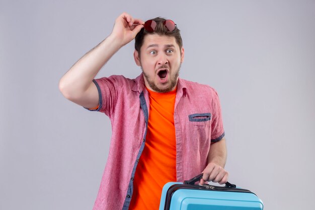 Młody przystojny podróżnik mężczyzna trzyma niebieską walizkę zaskoczony i zdumiony stojąc nad białą ścianą