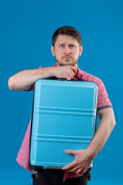 Młody przystojny podróżnik mężczyzna trzyma walizkę wyglądający na zdezorientowanego i bardzo niespokojnego stojącego nad niebieską ścianą