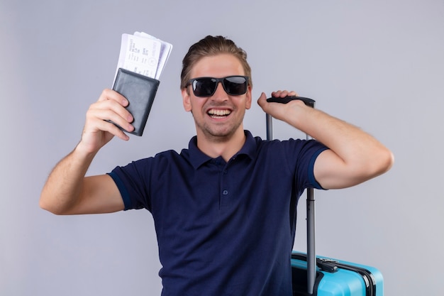 Młody przystojny podróżnik mężczyzna w czarnych okularach przeciwsłonecznych posiadający bilety lotnicze stojący z walizką szczęśliwy i wyszedł, uśmiechając się wesoło na białym tle