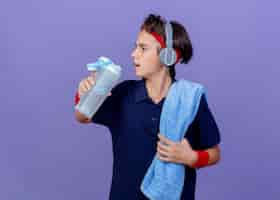 Bezpłatne zdjęcie młody przystojny sportowy chłopiec ubrany w opaskę na głowę, opaski na rękę i słuchawki z szelkami dentystycznymi i ręcznikiem na ramieniu, trzymając butelkę wody patrząc na bok odizolowany na fioletowej ścianie z miejscem na kopię