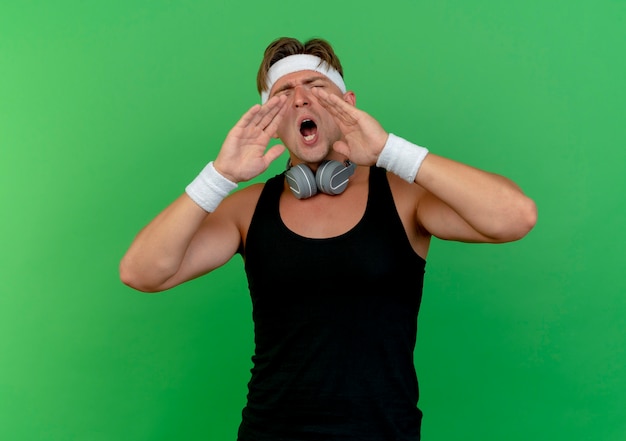 Bezpłatne zdjęcie młody przystojny sportowy mężczyzna nosi opaskę i opaski ze słuchawkami na szyi, kładąc ręce w pobliżu ust, wzywając kogoś na białym tle na zielonym tle