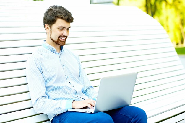 Młody przystojny uśmiechnięty biznesmena modela obsiadanie na parkowej ławce używać laptop w przypadkowym modnisia płótnie
