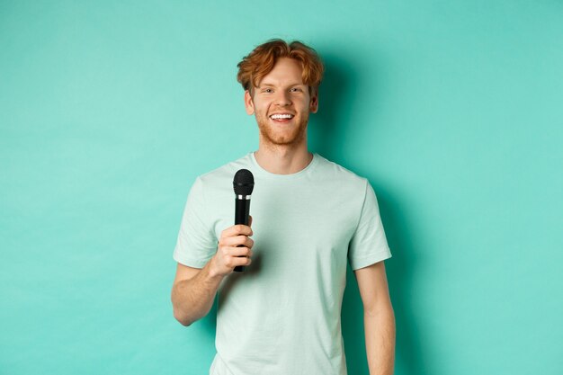 Młody rudy mężczyzna z brodą, ubrany w t-shirt, trzymający mikrofon i przemawiający, śpiewający karaoke, stojący nad miętowym tłem.