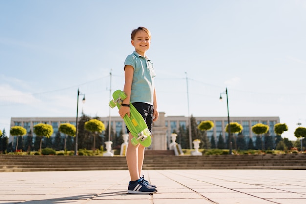 Młody SCHOOL fajny CHŁOPIEC w jasnych ubraniach, stojący z PENNY BOARD w rękach