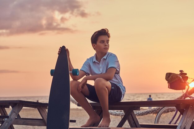 Młody skater chłopiec ubrany w t-shirt i szorty, siedząc na ławce na tle wybrzeża w jasny zachód słońca.