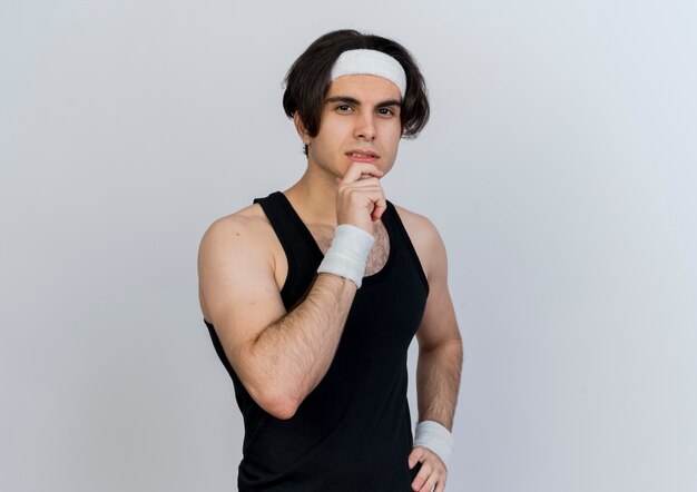 Młody sportowy człowiek ubrany w odzież sportową i opaskę patrząc z przodu ręką na brodzie myśli stojącej nad białą ścianą