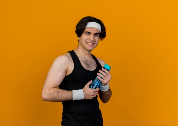 Młody sportowy człowiek ubrany w odzież sportową i pałąk trzymając butelkę wody, uśmiechając się