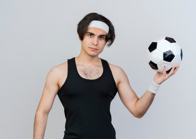 Młody sportowy człowiek ubrany w odzież sportową i pałąk trzymając piłkę nożną patrząc z przodu z poważną twarzą stojącą na białej ścianie