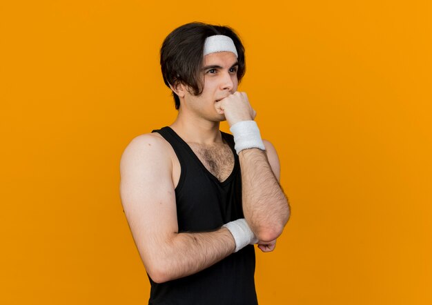 Młody sportowy człowiek ubrany w odzież sportową i pałąk ze słuchawkami na szyi, patrząc na bok z ręką na brodzie myśli
