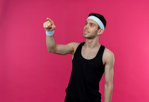 Młody sportowy mężczyzna w opasce patrząc na bok, wskazując palcem wskazującym na coś uśmiechniętego stojącego na różowej ścianie