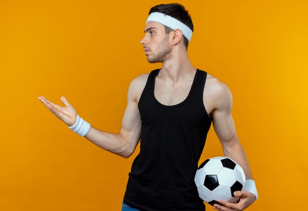 Młody sportowy mężczyzna w opasce trzyma piłkę nożną patrząc na bok z wyciągniętym ramieniem, pytając lub argumentując stojąc nad pomarańczową ścianą
