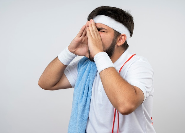 Młody sportowy mężczyzna z zamkniętymi oczami na sobie opaskę i opaskę z ręcznikiem i skakankę na ramieniu wzywając kogoś na białej ścianie