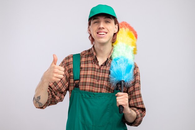 Młody sprzątacz w kombinezonie w kratę koszulę i czapce, trzymający kolorową miotełkę, uśmiechający się radośnie pokazując kciuk do góry stojący nad białą ścianą