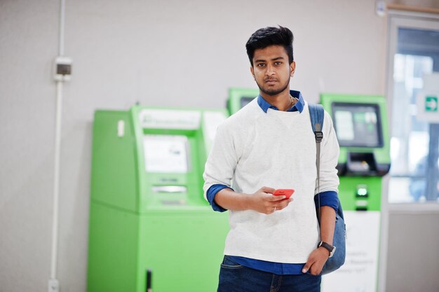 Młody stylowy azjatycki mężczyzna z telefonem komórkowym i plecakiem przed rzędem zielonego bankomatu