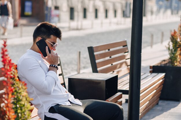 Młody stylowy facet w koszuli z telefonem na ławce w słoneczny dzień na zewnątrz