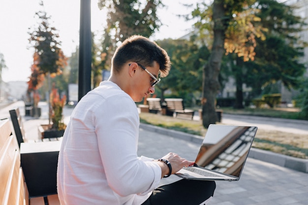 Młody stylowy facet w koszuli z telefonu i notebooka na ławce w słoneczny ciepły dzień na zewnątrz, niezależny