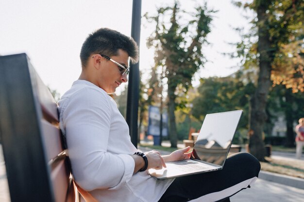 Młody stylowy facet w koszuli z telefonu i notebooka na ławce w słoneczny ciepły dzień na zewnątrz, niezależny